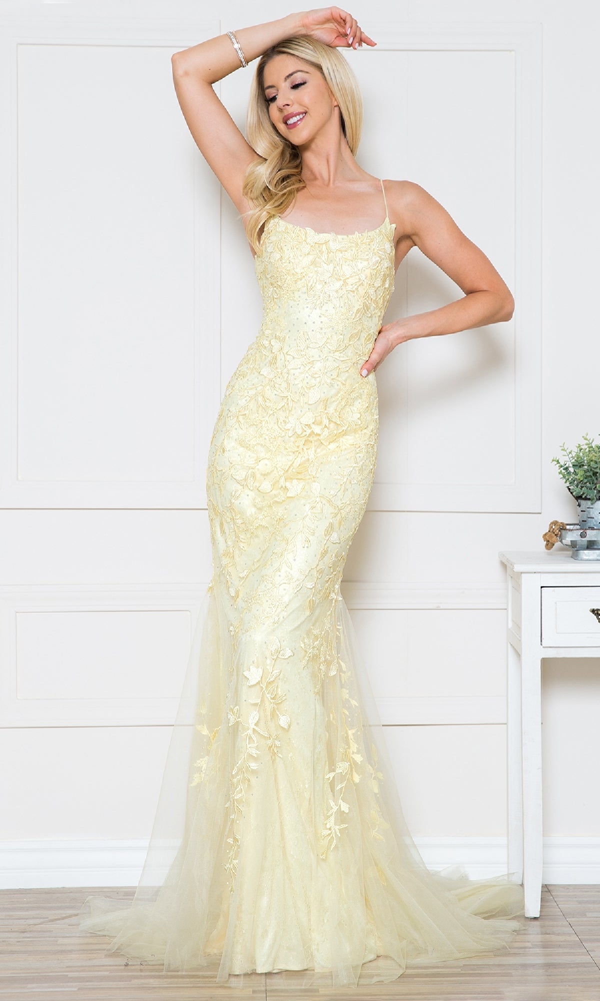 A woman stands indoors, smiling and posing with one arm raised, wearing the Embroidered-Lace Tight Long Prom Gown 799—a pale yellow evening dress with thin straps.
